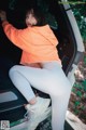 A woman in an orange sweatshirt and white leggings sitting in the back of a car.