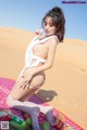 A woman in a white bikini sitting on a blanket in the desert.